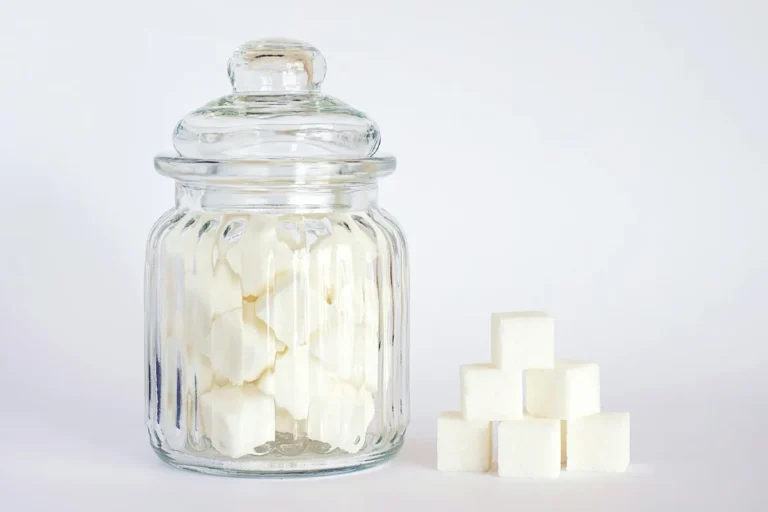 Close-up of sugar cubes stacked inside and beside a clear glass jar against a white background